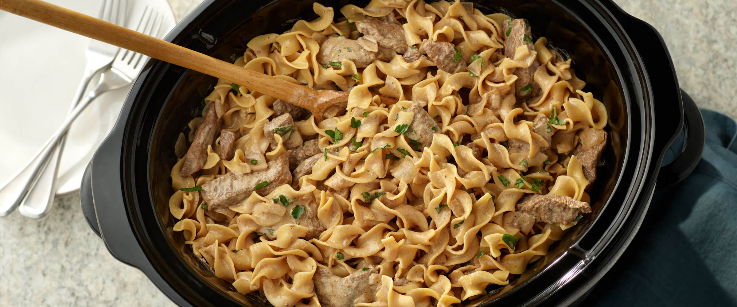 Slow Cooker Beef and Noodles in black pot with wooded serving spoon