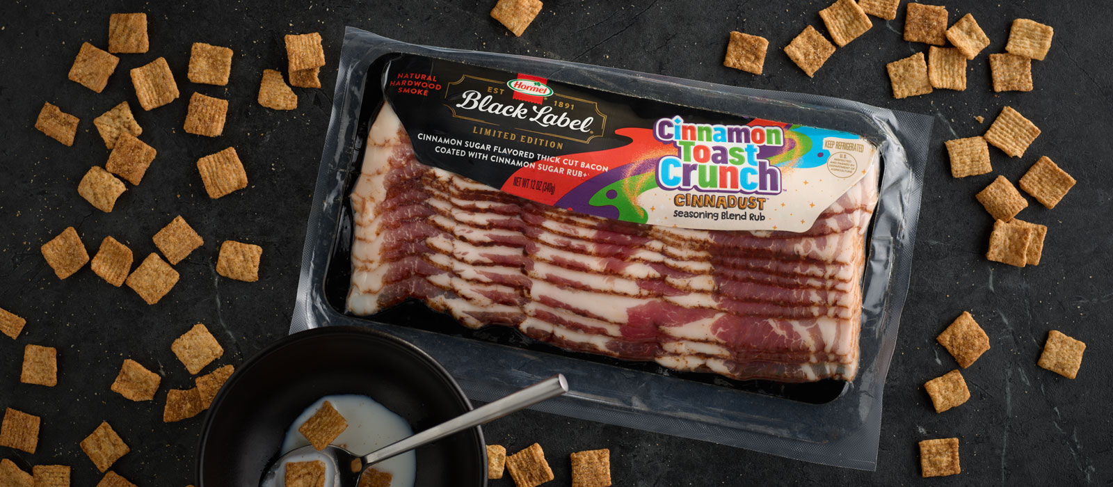 Packaged Hormel Black Label Cinnamon Toast Crunch flavored bacon on a dark surface, surrounded by scattered pieces of Cinnamon Toast Crunch cereal and a small bowl with milk.