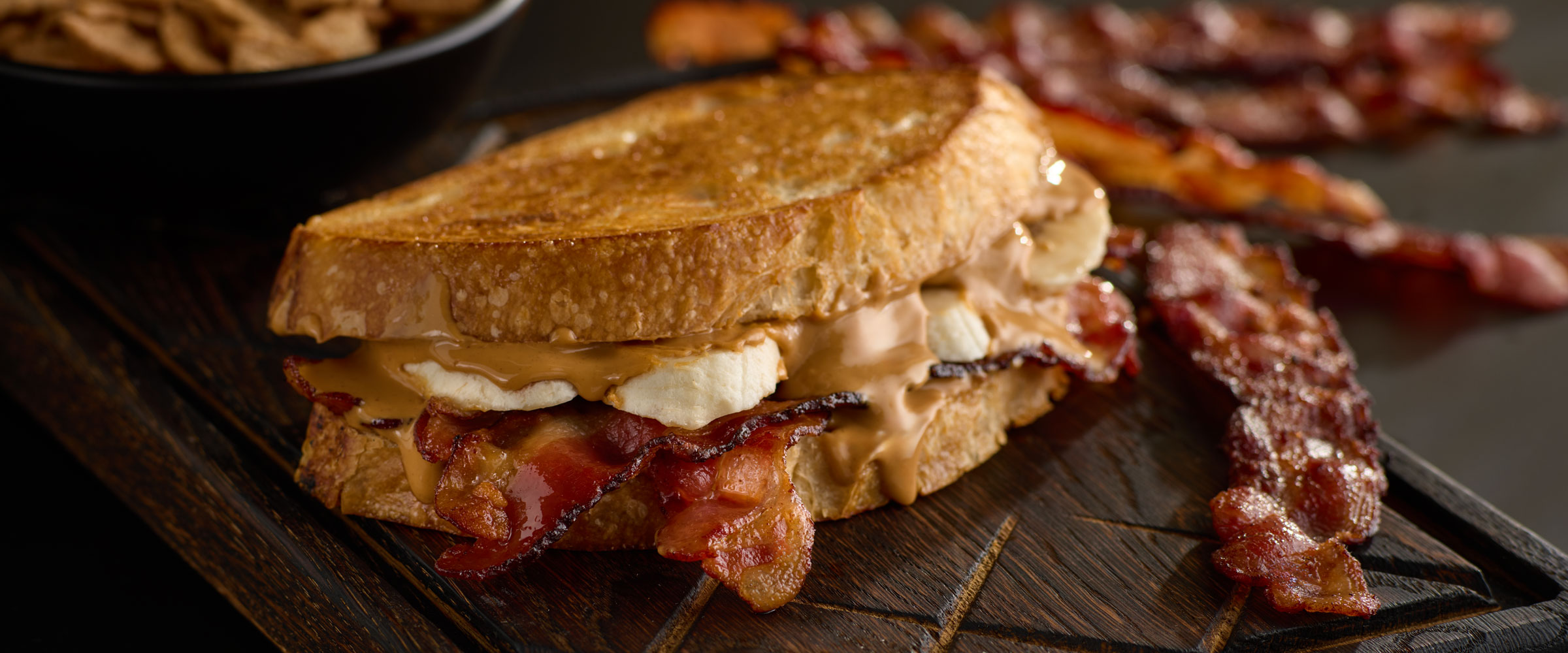 Close-up of a grilled sandwich with bacon, melted cheese, and chicken on a wooden cutting board, accompanied by crispy bacon strips and chips in the background.