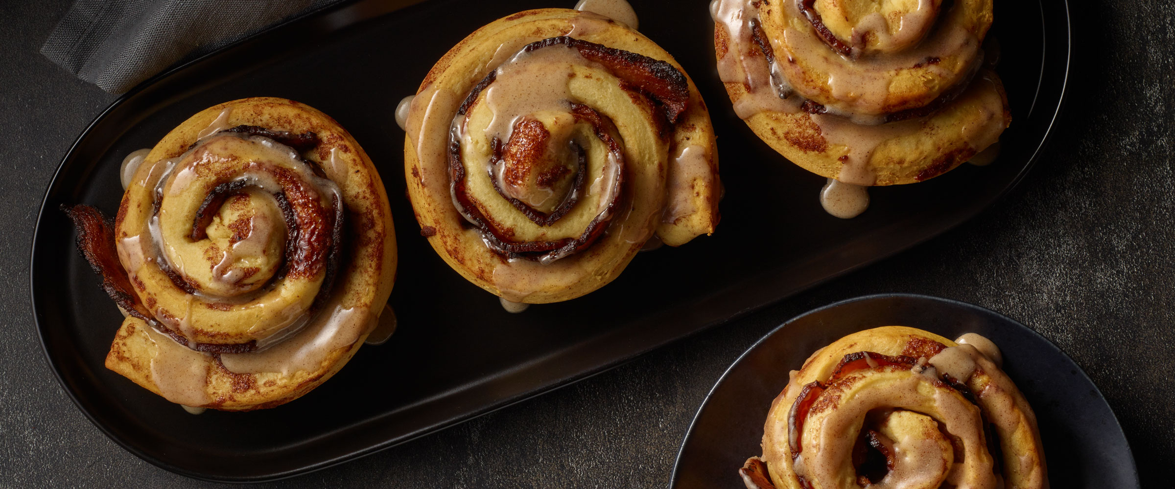 Four cinnamon rolls with a strip of bacon inside topped with icing on a dark oval plate, set against a dimly lit background.