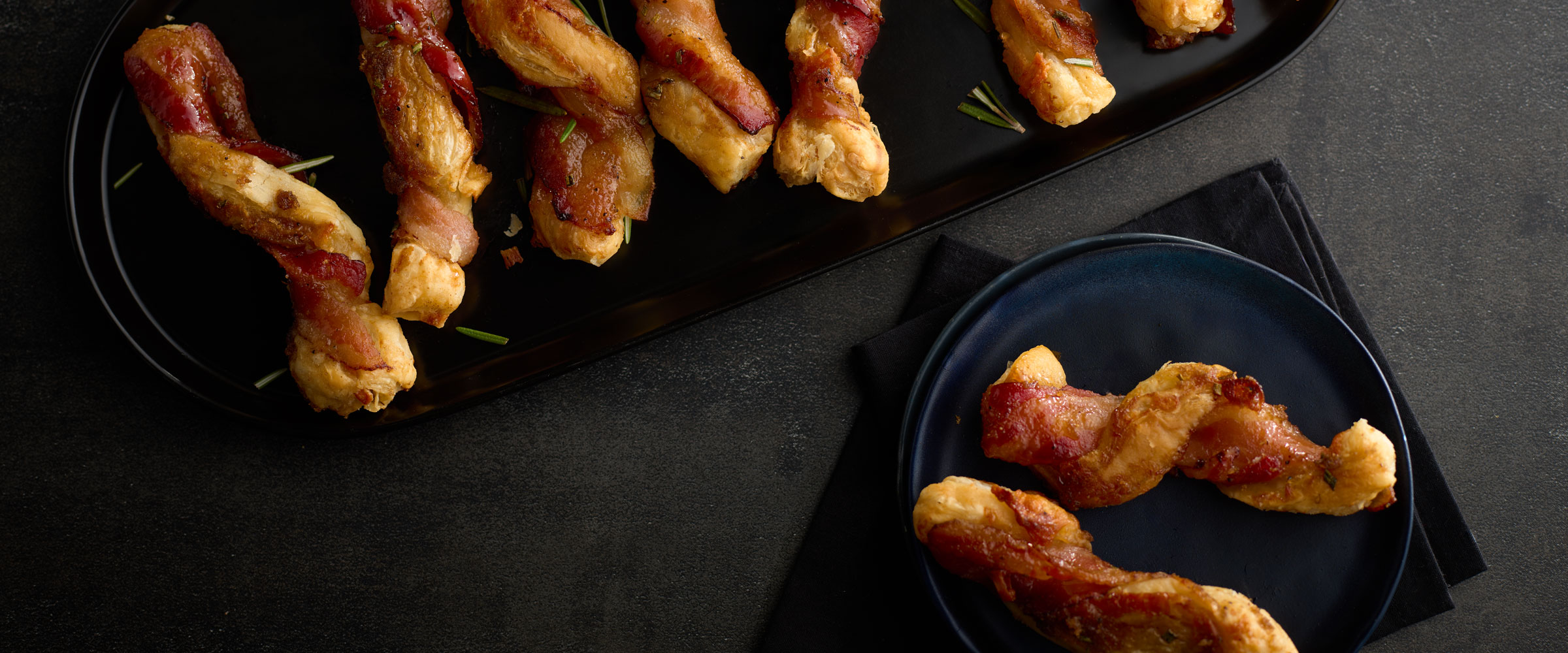 A selection of pastries, wrapped with crispy bacon strips served on a sleek black plate and a dark textured background.