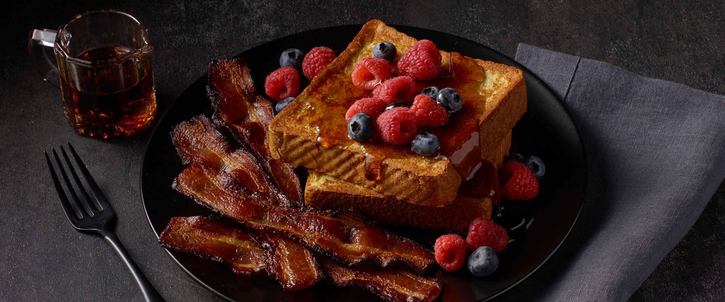 A plate of French toast topped with raspberries and blueberries, accompanied by crispy bacon strips, with a glass of maple syrup on the side. All set on a dark, textured surface.