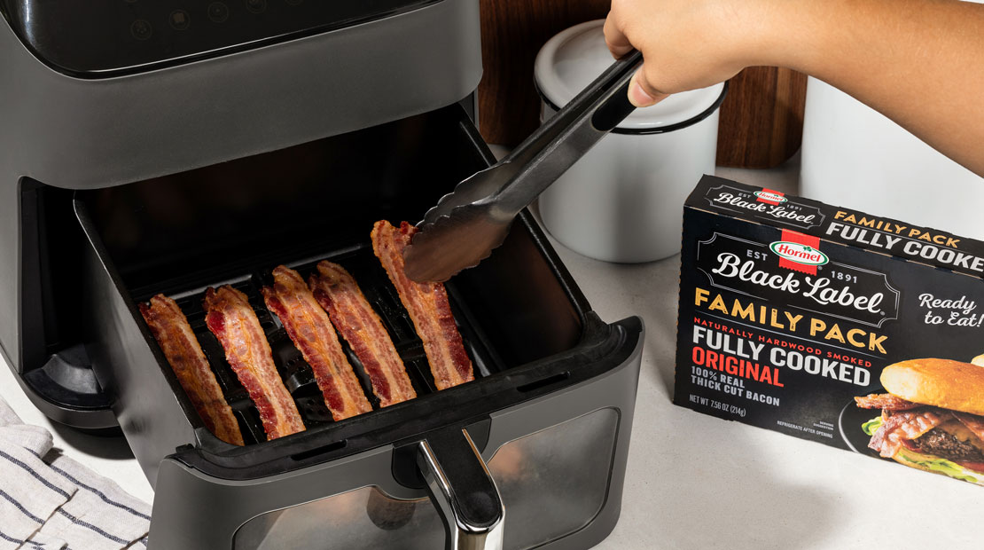 Person using tongs to remove bacon strips from a black air fryer, with a package of HORMEL® BLACK LABEL® Fully Cooked Original Bacon’ visible on the countertop.