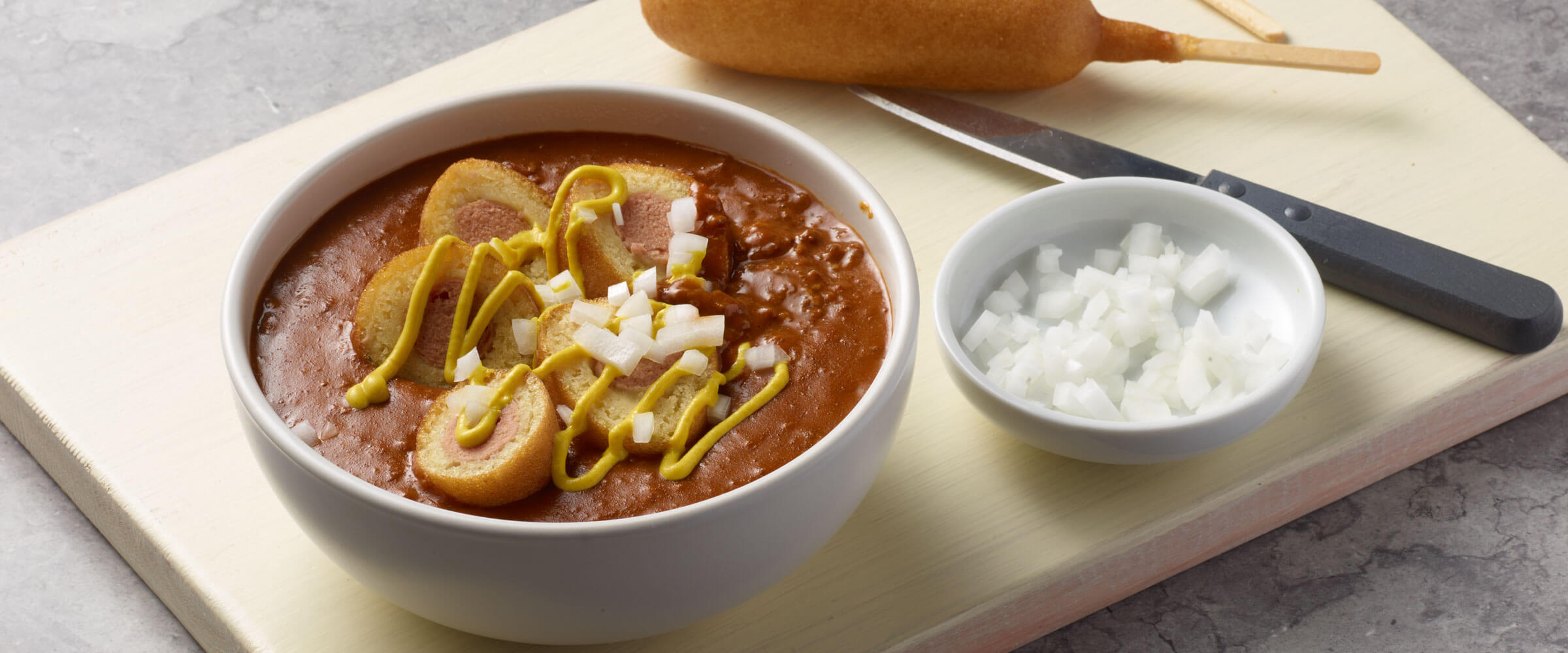 Chili topped with Corndogs slices, mustard and onions in white bowl with extra onions on the side