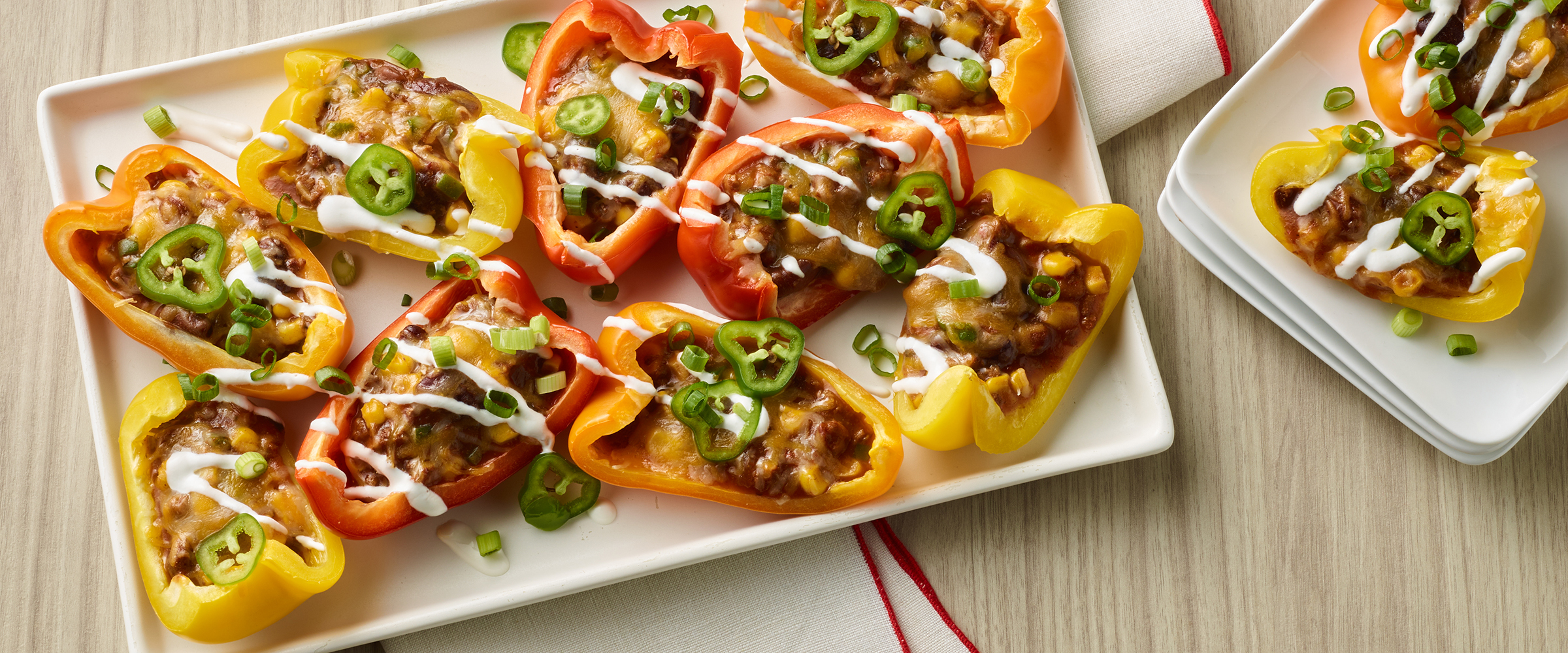 A rectangular plate displaying stuffed bell peppers in orange and yellow, topped with melted cheese, chili and sliced green jalapeños.