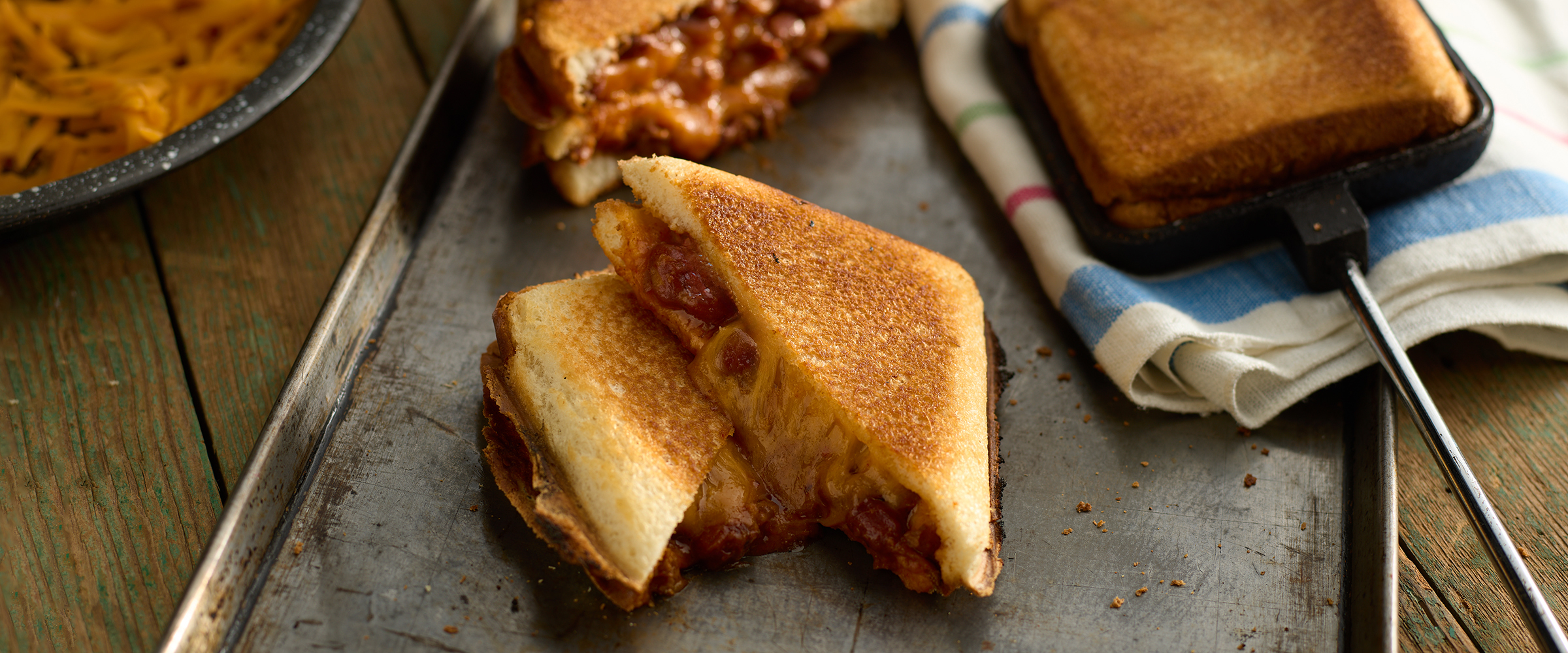 A grilled cheese sandwich cut in half, revealing melted cheese and chili, served on a metal tray with two whole sandwiches in the background on a wooden table.