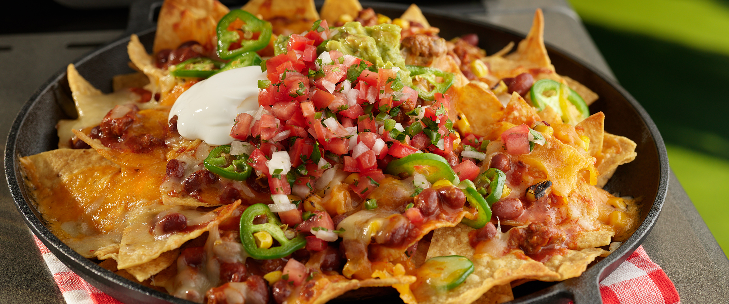 A platter of nachos generously topped with melted cheese, sliced jalapeños, diced tomatoes, chunks of avocado, and a dollop of sour cream. The nachos are spread out on a round black plate, which sits on a dark countertop.