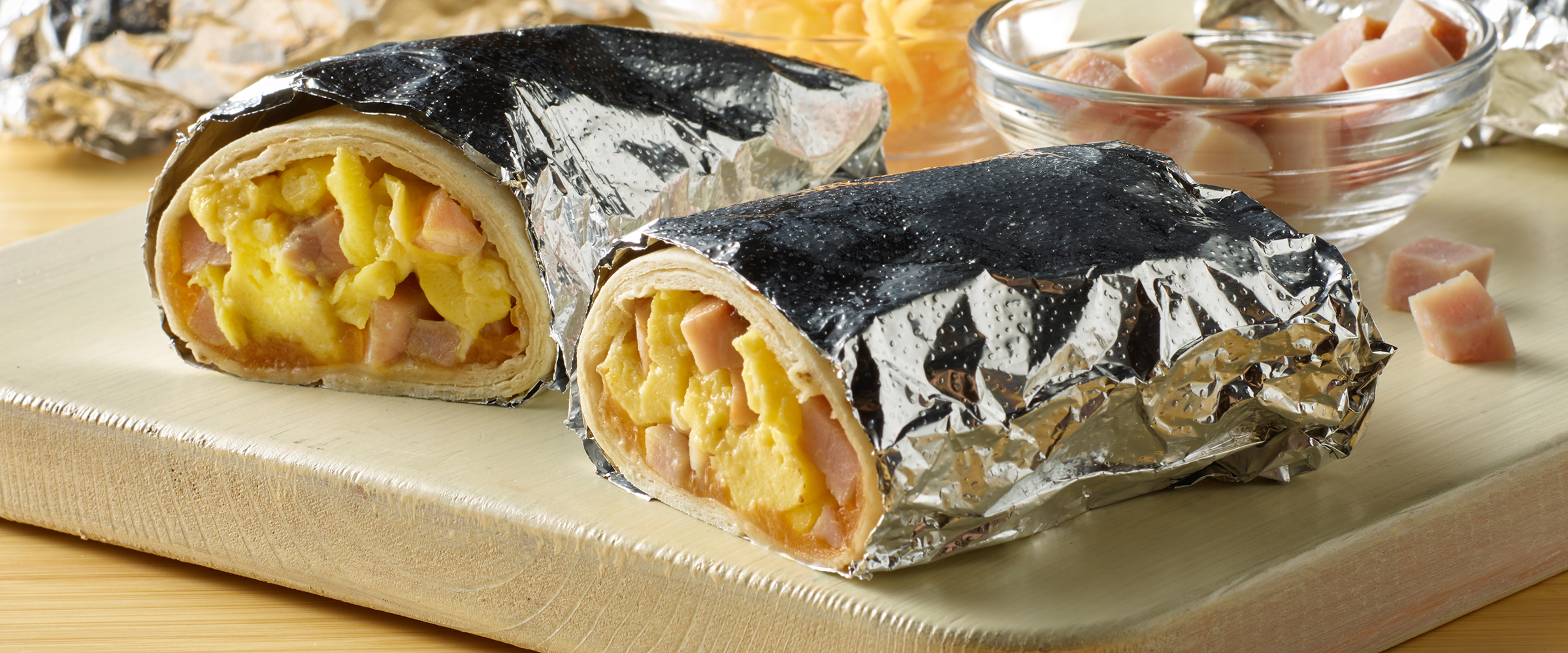 A breakfast burrito wrapped in foil with one end cut open to reveal scrambled eggs, cheese, and cubes of ham, placed on a wooden serving board alongside a small bowl of salsa.