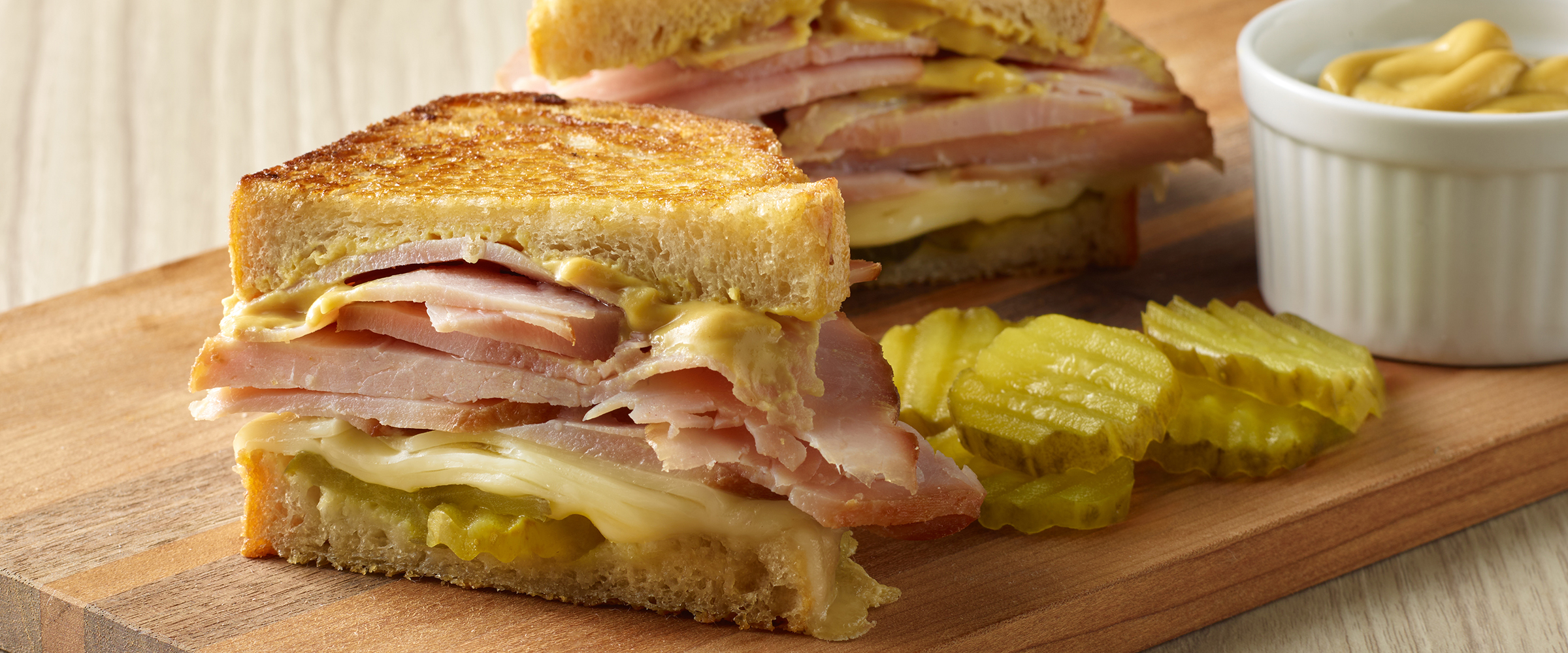 A grilled ham and cheese sandwich on a wooden cutting board, accompanied by pickles and a small bowl of mustard.