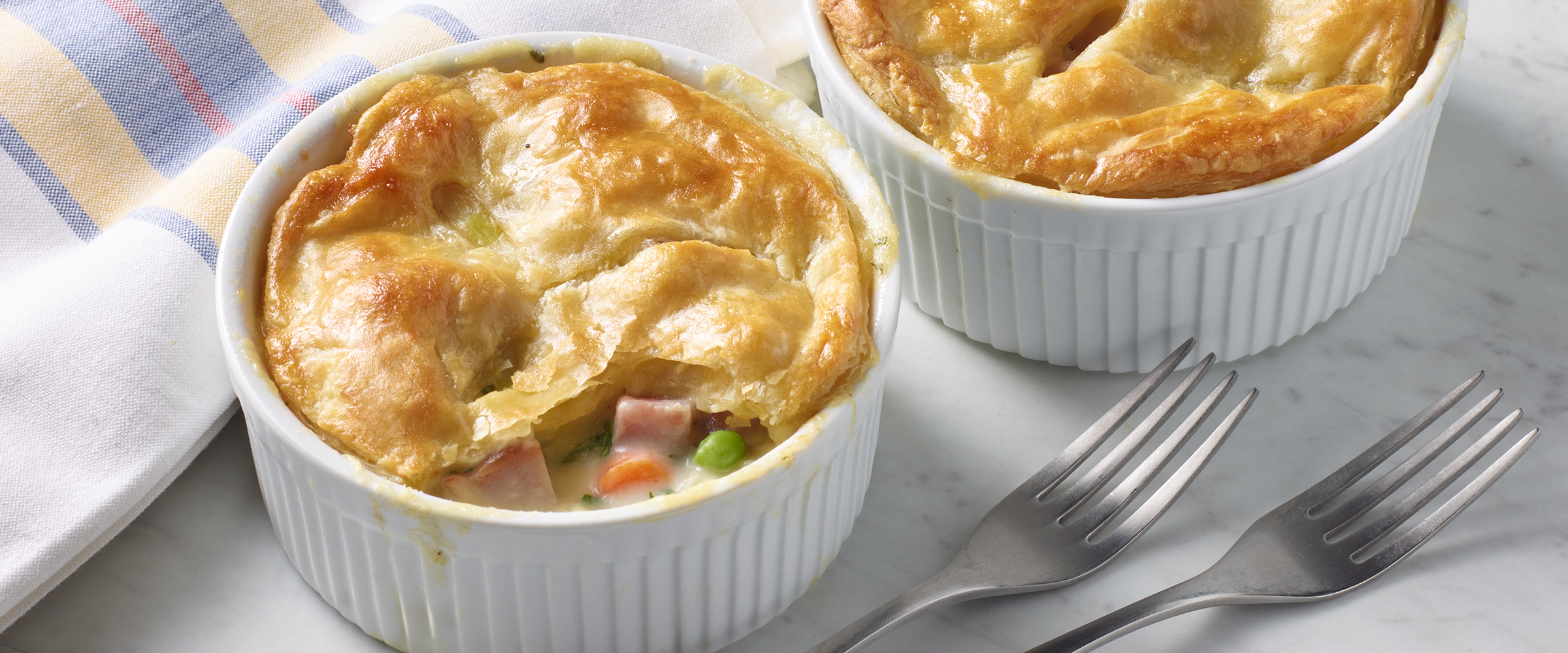 Two chicken pot pies in white ramekins on a marble countertop, one with a golden-brown crust broken open to reveal filling with visible pieces of chicken, ham, and peas, served with two forks beside them.