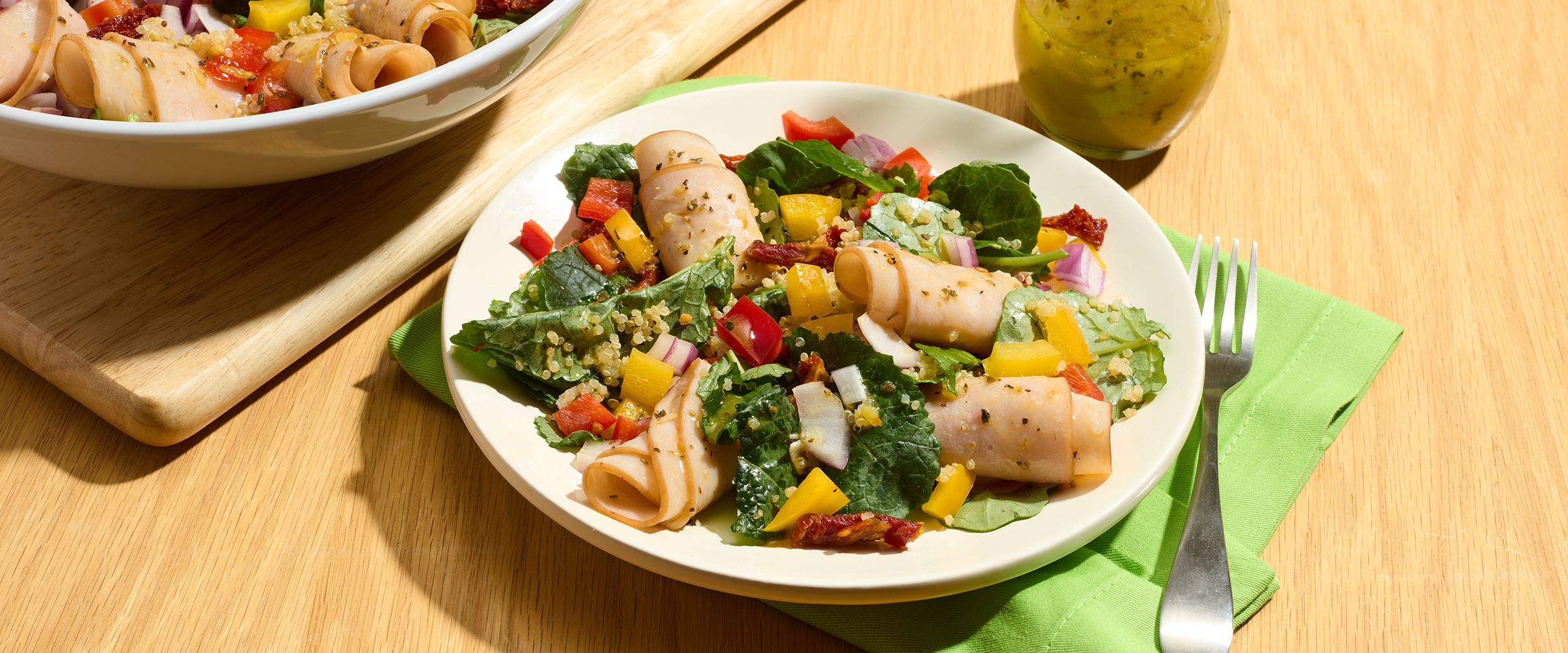 Two plates filled with colorful salads on a wooden table. The salad on the left features pasta, tomatoes, and greens, and the one on the right includes mixed greens, multi-colored bell peppers, and cubes of mango, sprinkled with seeds. A green napkin underlies the plates, and a pear is visible in the background.