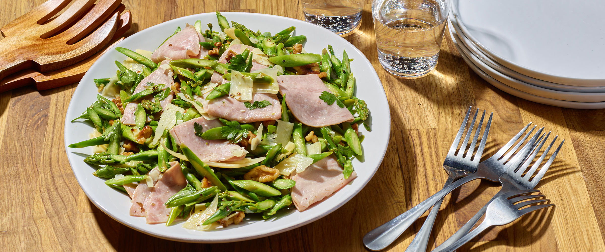 A rectangular platter of green bean salad with sliced ham, almonds, and garnished with herbs, accompanied by two forks on a wooden dining table with empty plates and water glasses nearby.