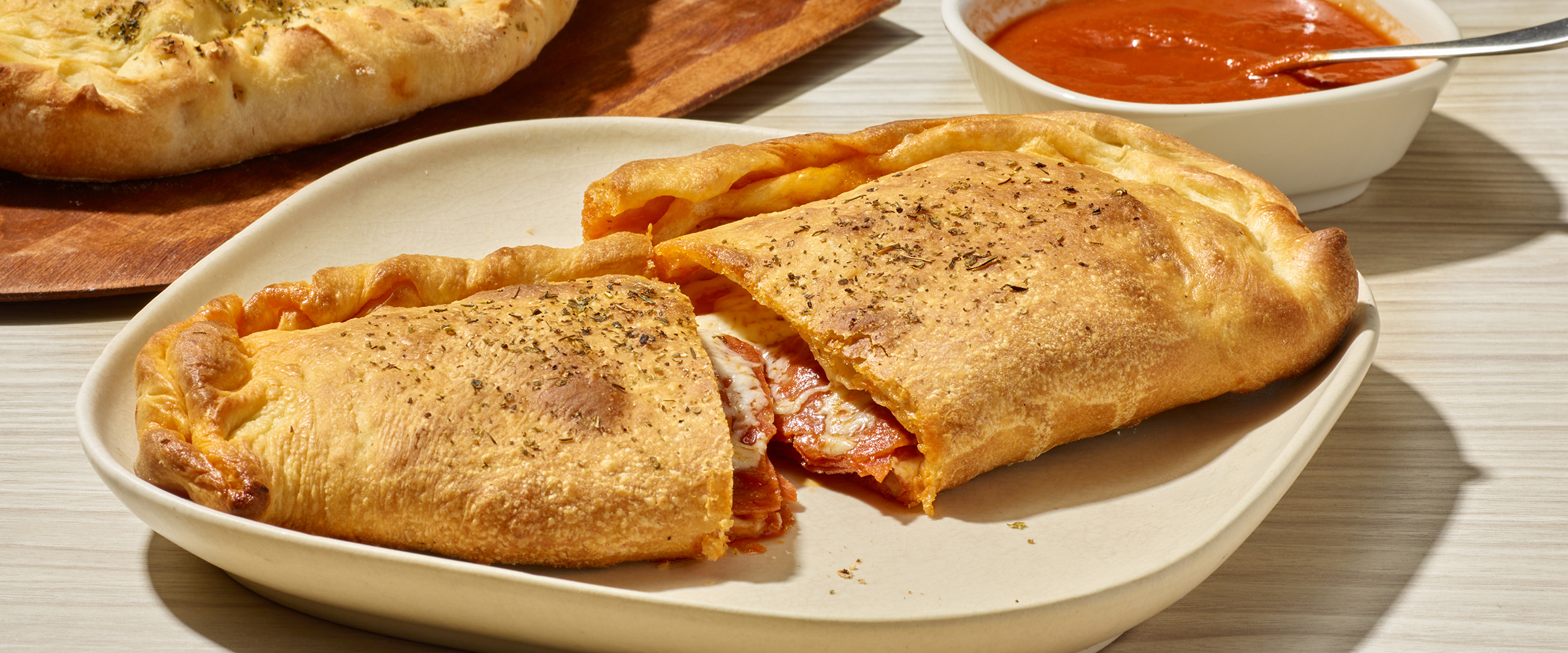 Two freshly baked calzones on a plate, one sliced open revealing a hot, cheesy filling, accompanied by a bowl of marinara sauce on a wooden table.