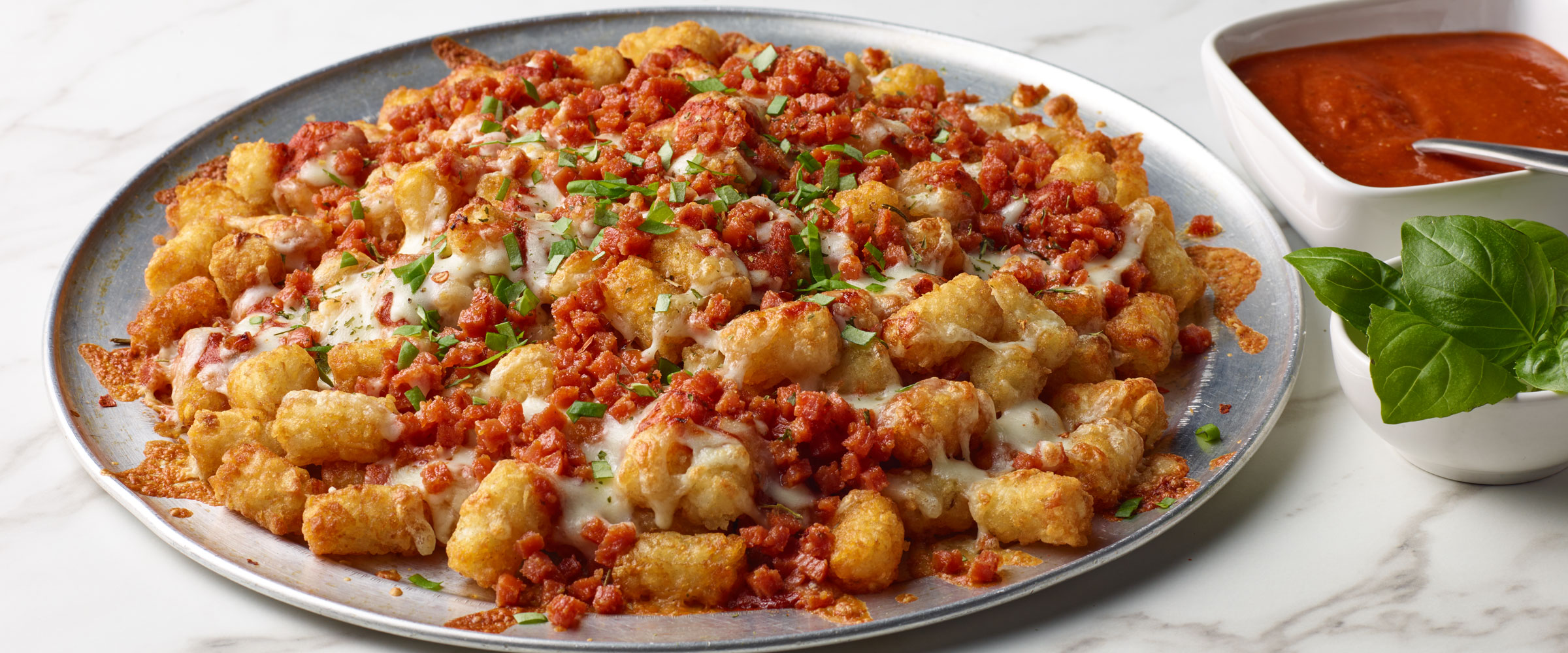 A large pizza topped with golden tater tots, melted cheese, crispy diced pepperoni, and chopped herbs, served with a side of marinara sauce.