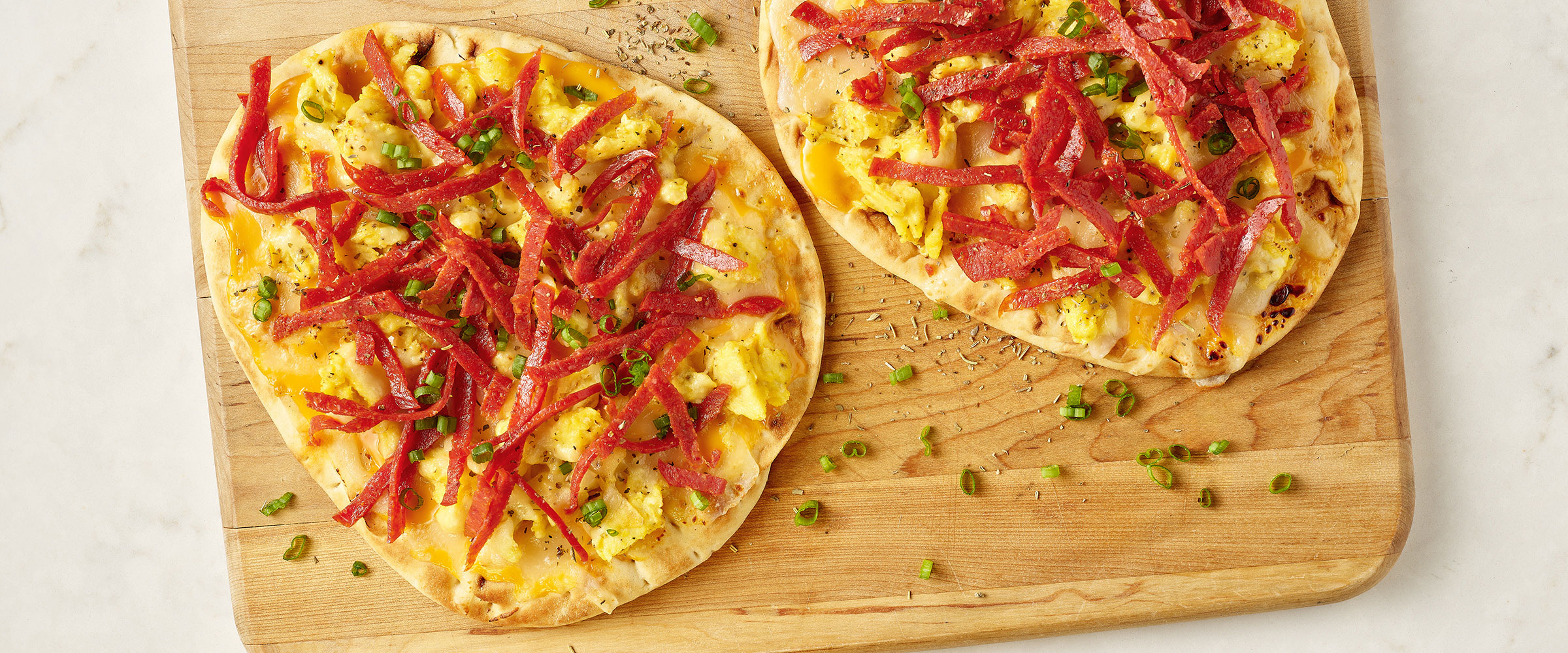 Flatbread pizzas topped with yellow cheese, scrambled eggs, green herbs, and confetti pepperoni, served on a wooden cutting board.