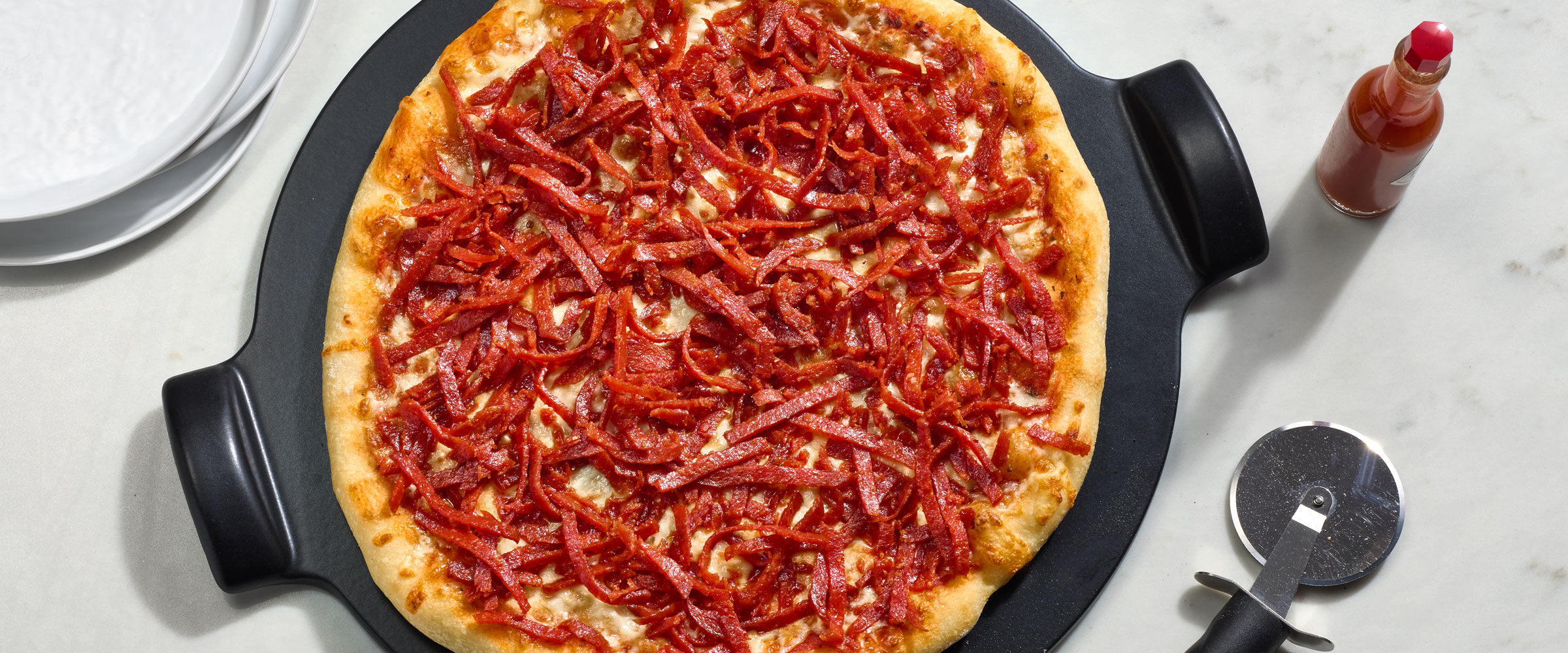 A pizza with a generous topping of pepperoni on a black serving tray, accompanied by a pizza cutter and a bottle of hot sauce.