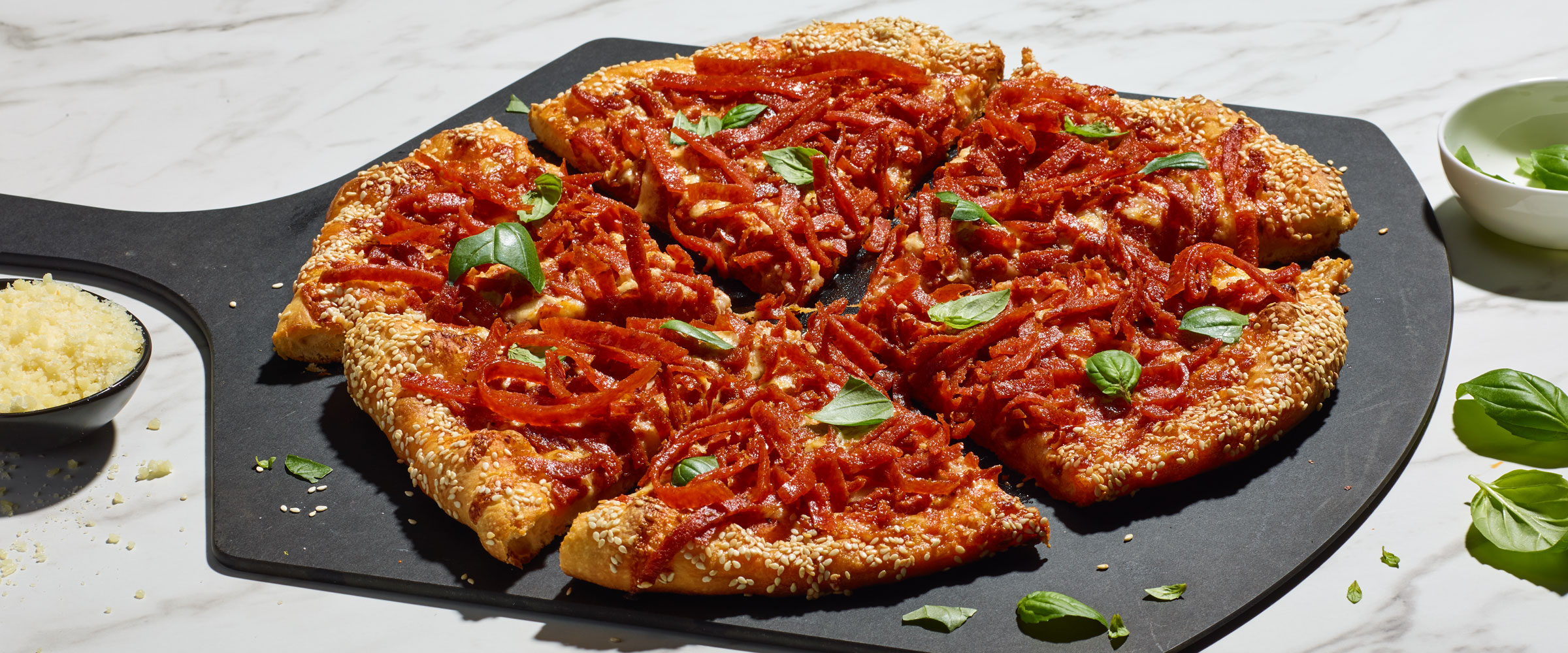 A whole pepperoni pizza with fresh basil on top, served on a black cutting board, with a small bowl of grated cheese and a dish of green basil leaves on the side.