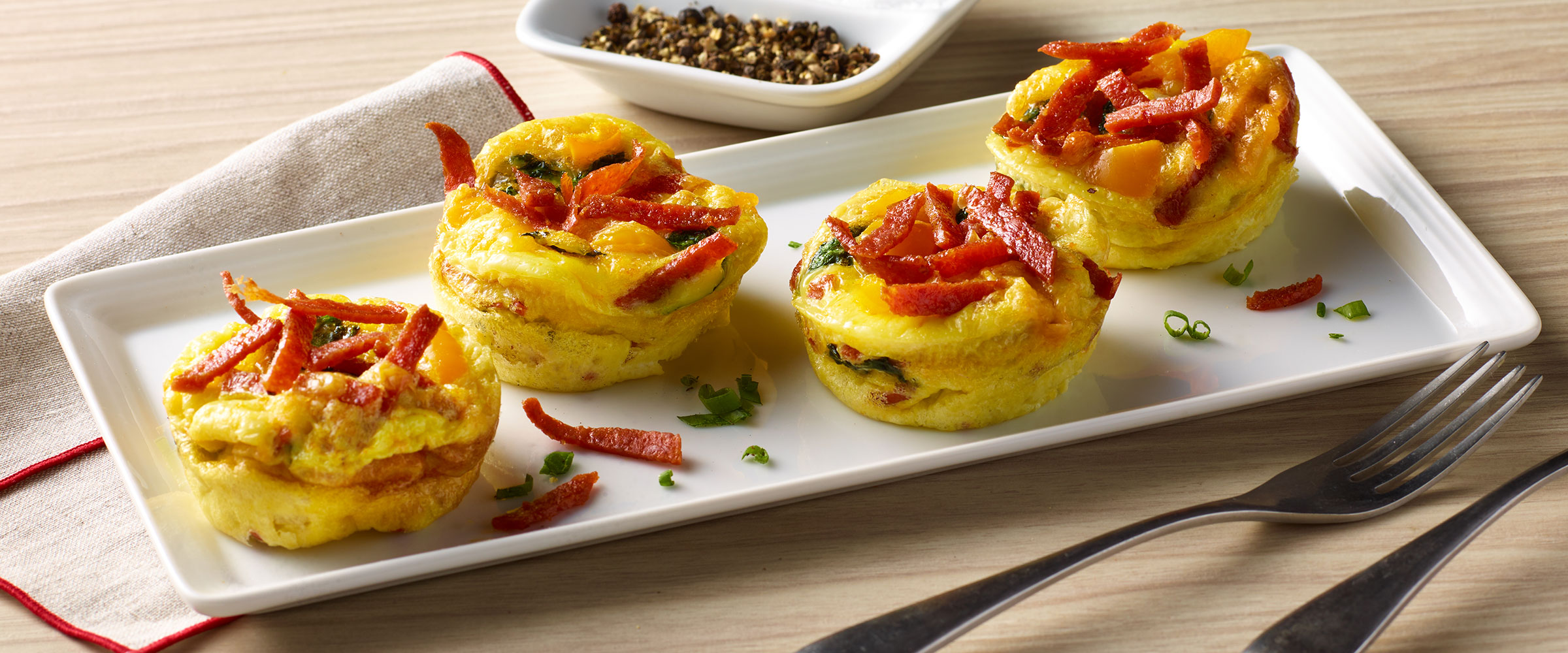 Three egg muffins topped with confetti pepperoni and herbs, served on a white rectangular plate with a side of red peppers and seasonings, presented on a wooden table.