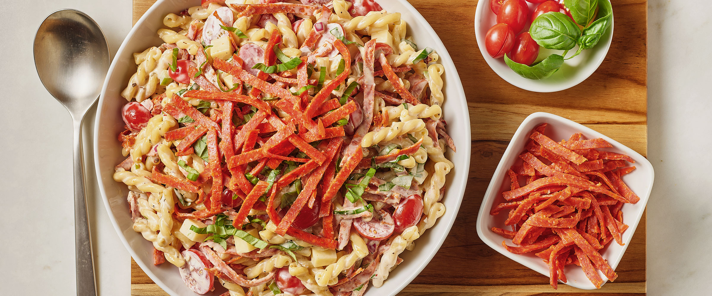 A bowl of pasta salad with sliced red peppers, pepperoni, and fresh basil, accompanied by a small bowl of additional confetti pepperoni strips on the side.