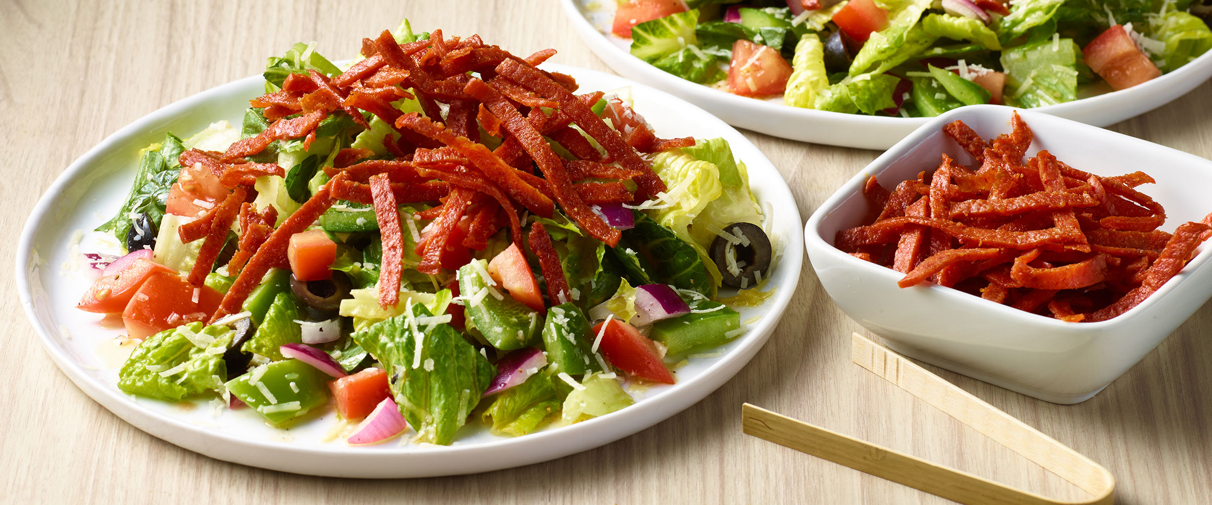 A plates of salad containing lettuce, tomatoes, cucumber, onions, and bell peppers, topped with a generous amount of confetti pepperoni strips, served alongside an additional bowl of bacon strips.