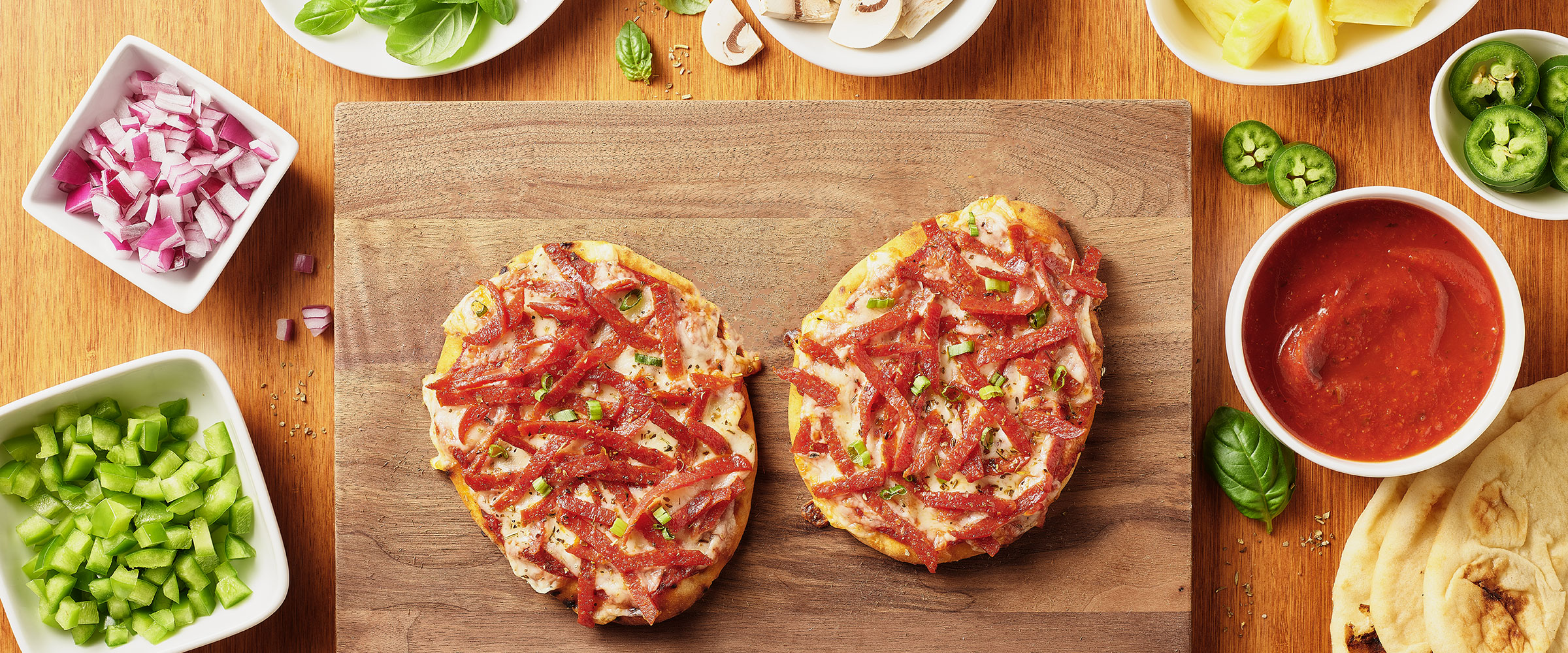 Wooden table with two pepperoni flatbreads and assorted ingredients like basil, onions, jalapenos, tomato sauce, and mushrooms around them.