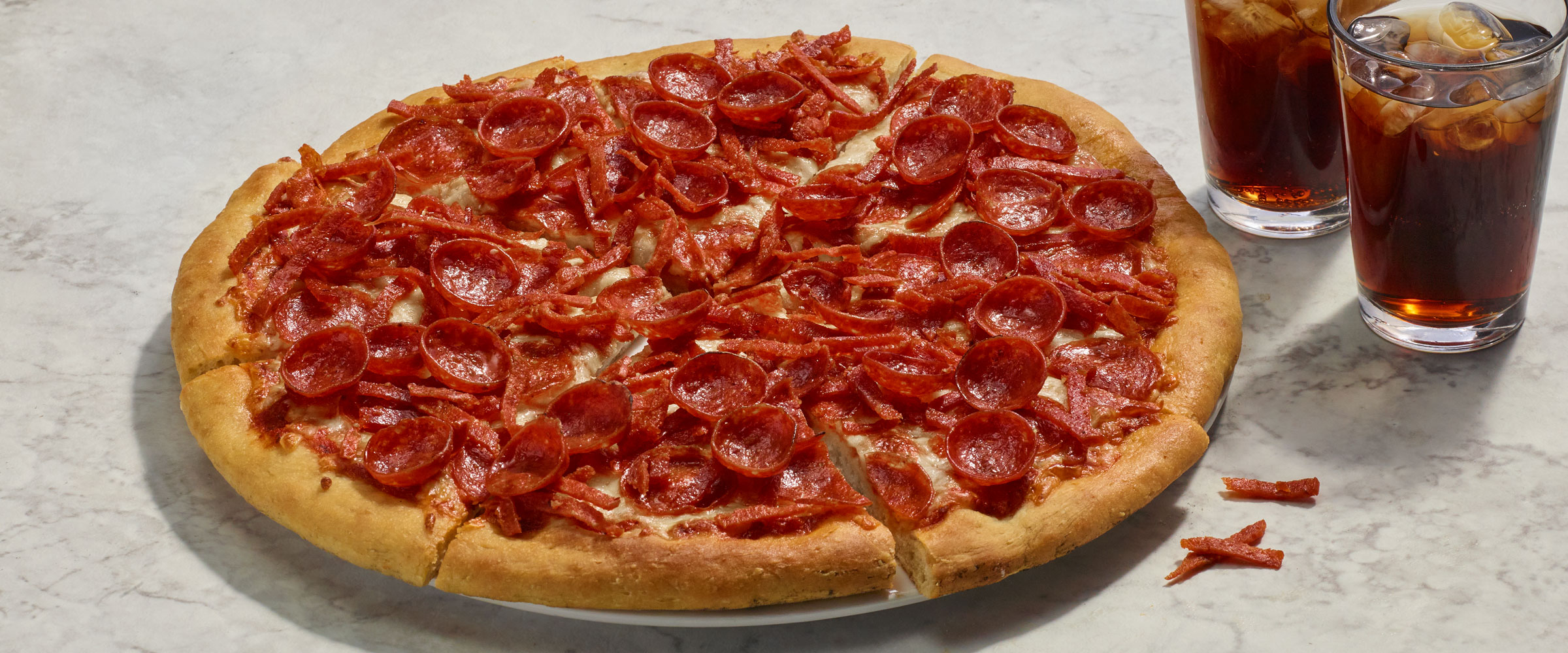 A large pepperoni pizza, with strips of pepperoni, cupped pepperoni and regular slices on a light-colored countertop, accompanied by two glasses of soda with ice.