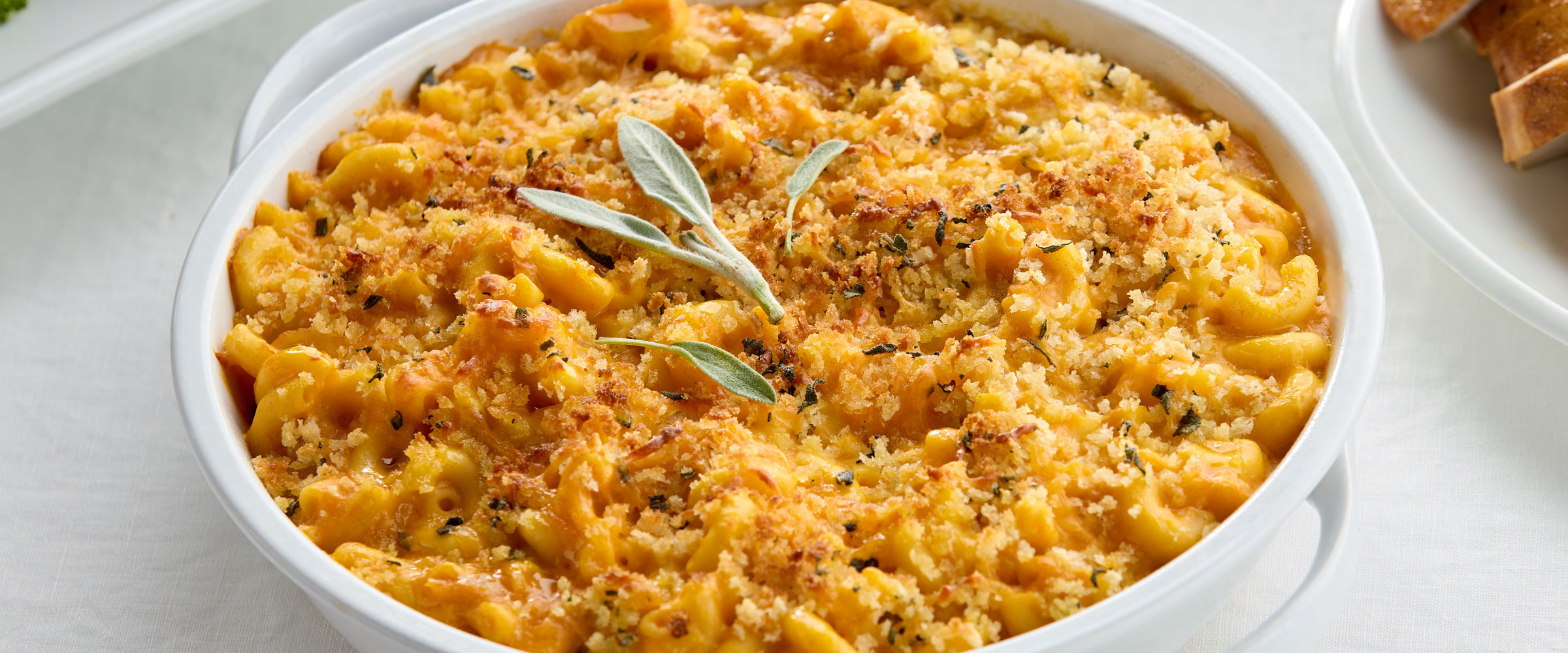 A dish of baked macaroni and cheese topped with breadcrumbs and garnished with sage leaves, served in a white oval dish next to slices of bread on a table.